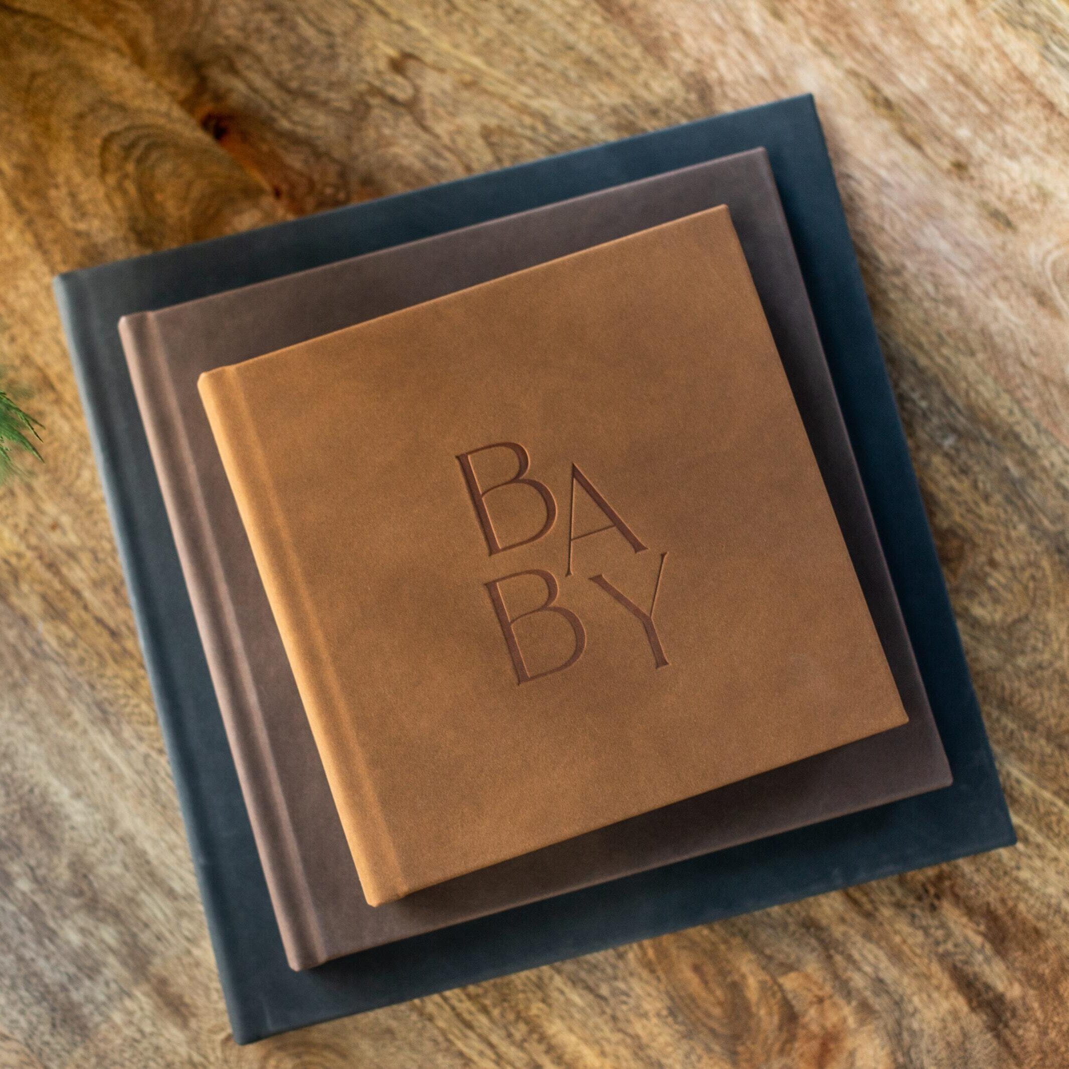 Stacked professional photo albums in multiple sizes debossed with a custom BABY die on a vintage camel leather cover, displayed on a rustic wood surface.