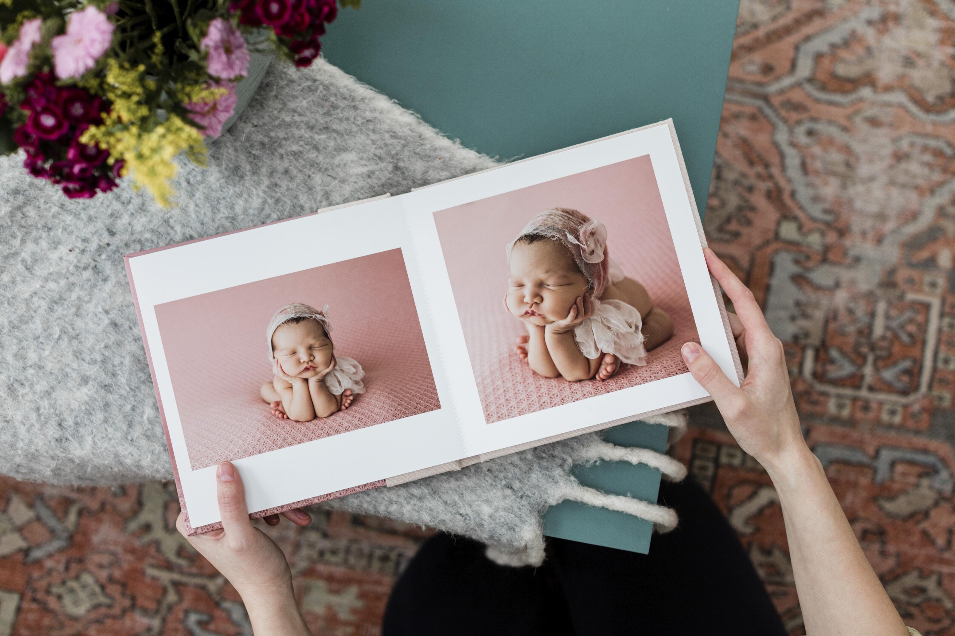 Open baby photo album with lay-flat binding showing newborn portrait spreads printed on archival photographic paper, held in a mother's hands.