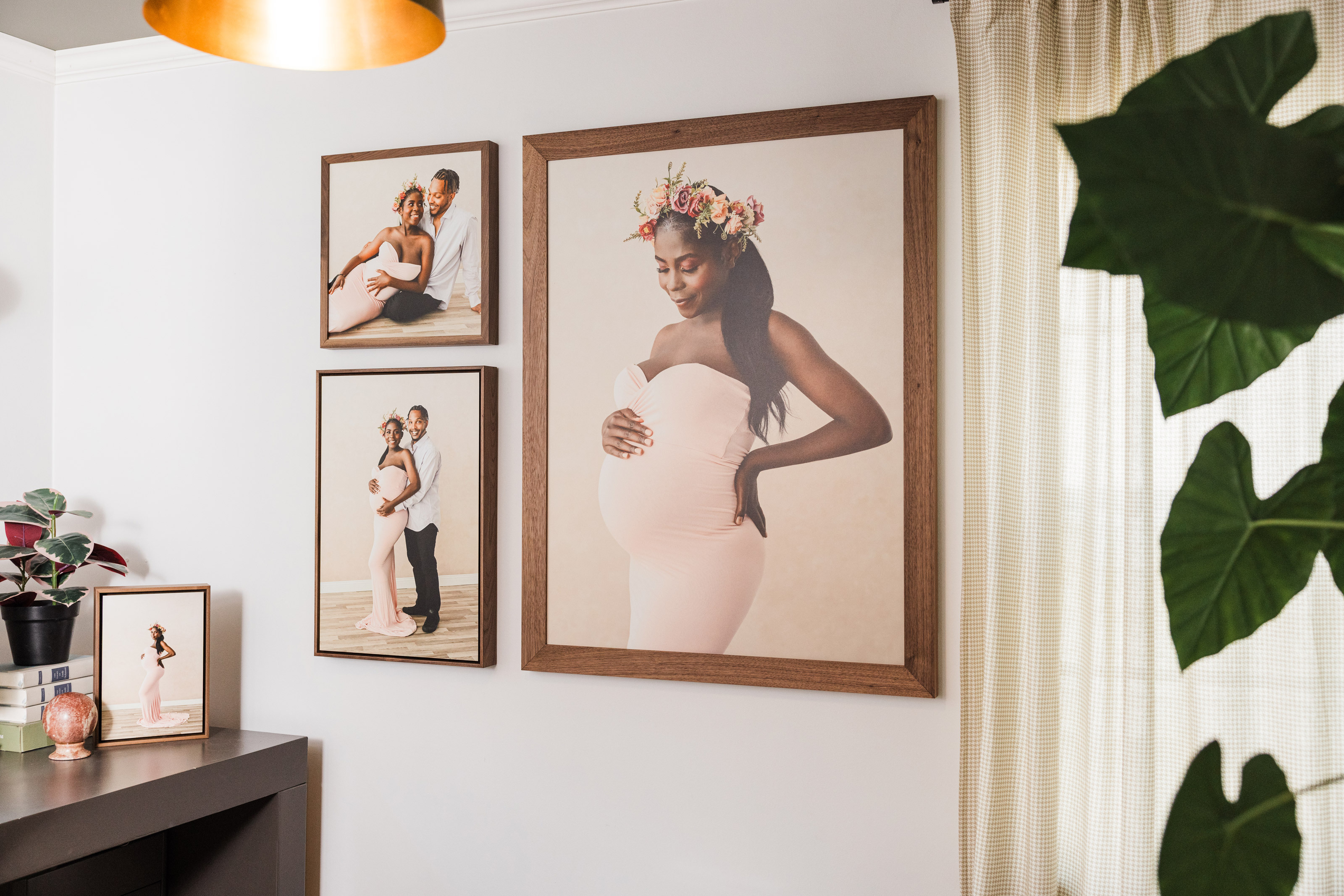 Wall collage of three maternity session prints framed with walnut wood mouldings on a home office wall, plant in foreground with desk in the background