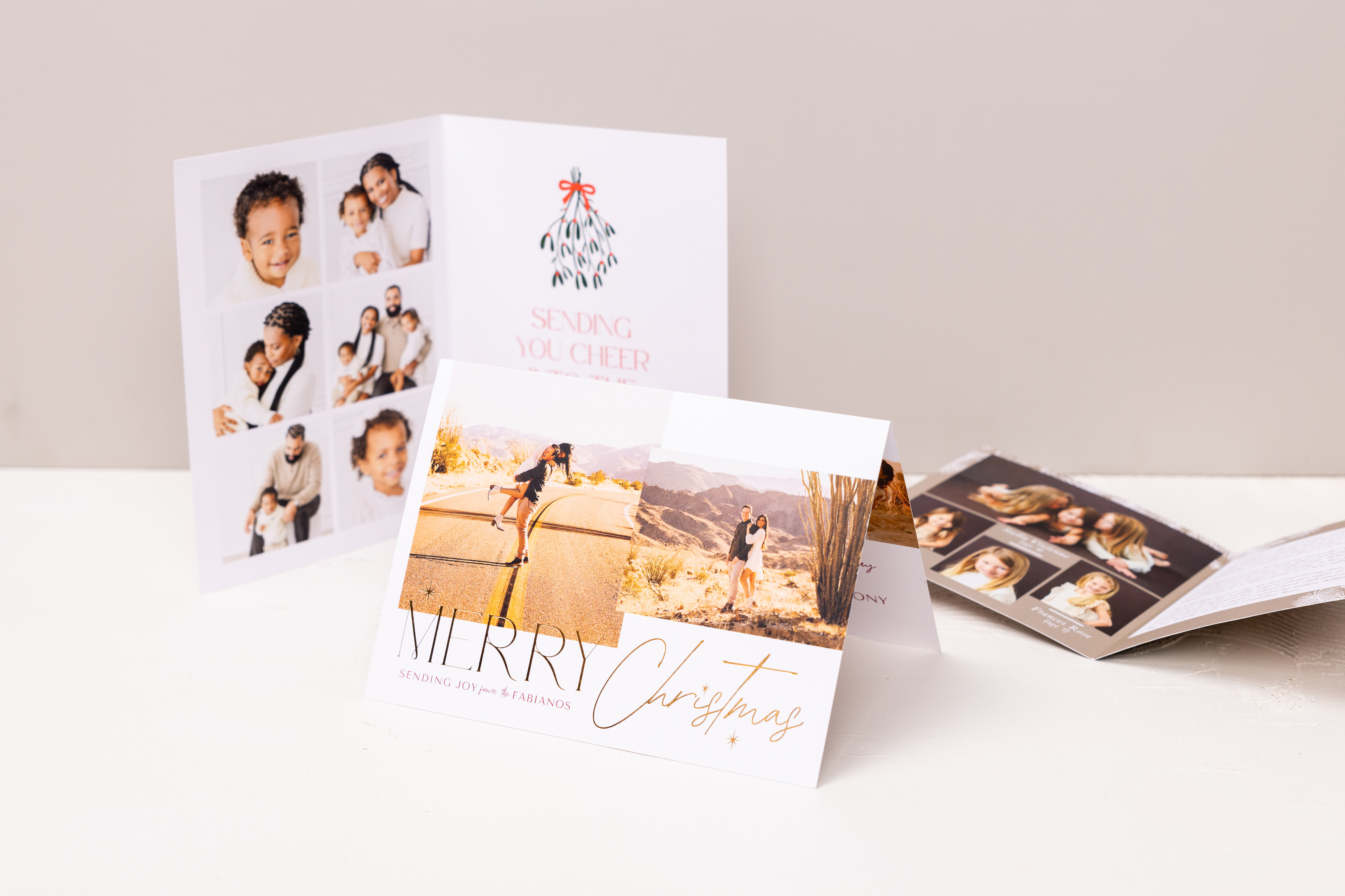 Three Folded Cards on a white surface with tan background, central card features desert wedding portraits on a 5×7 Folded Card with gold foil-pressed "Merry Christmas" design