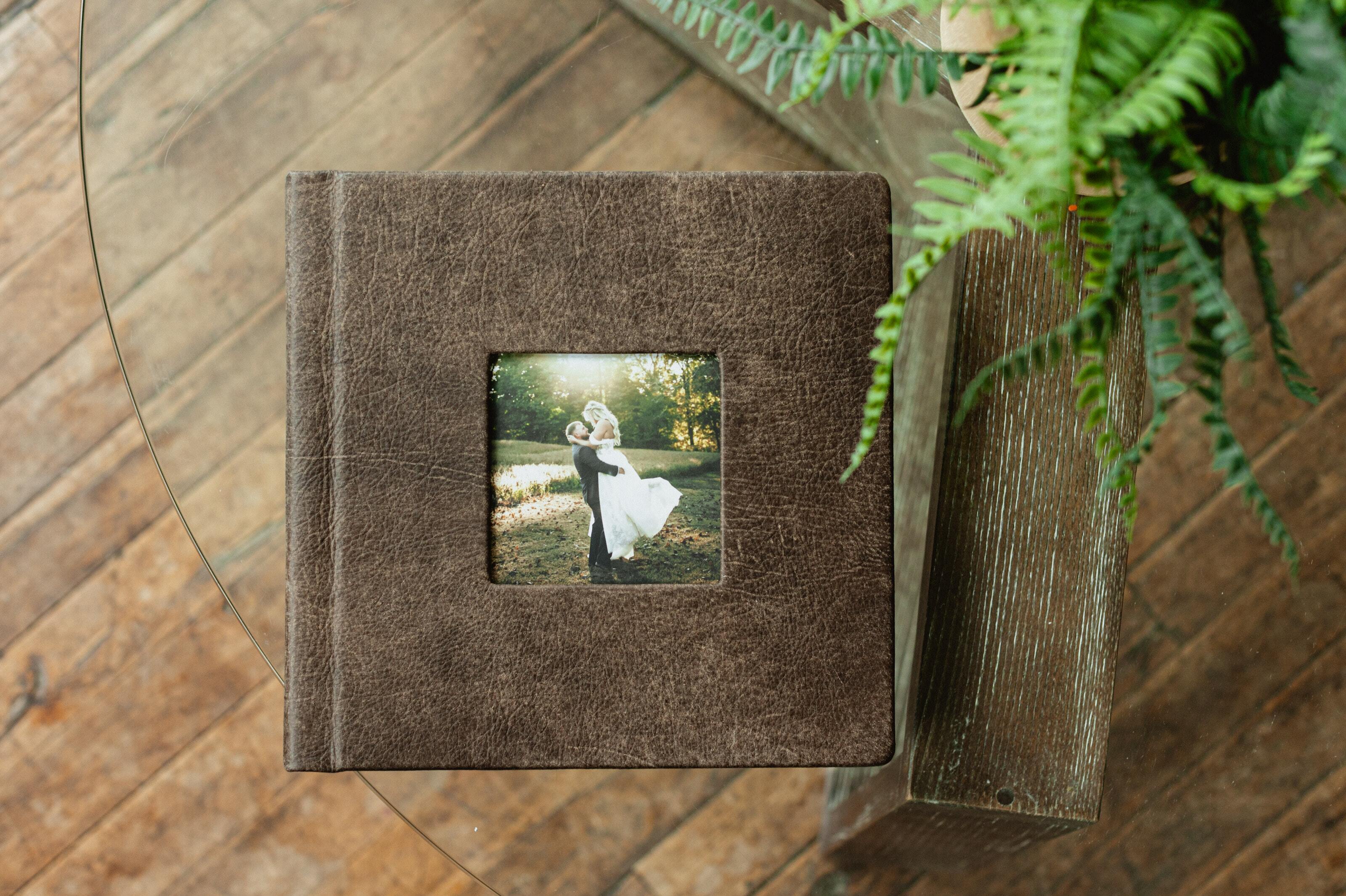 Professional wedding photo album with distressed leather cameo cover displaying a bride and groom portrait on a glass table.
