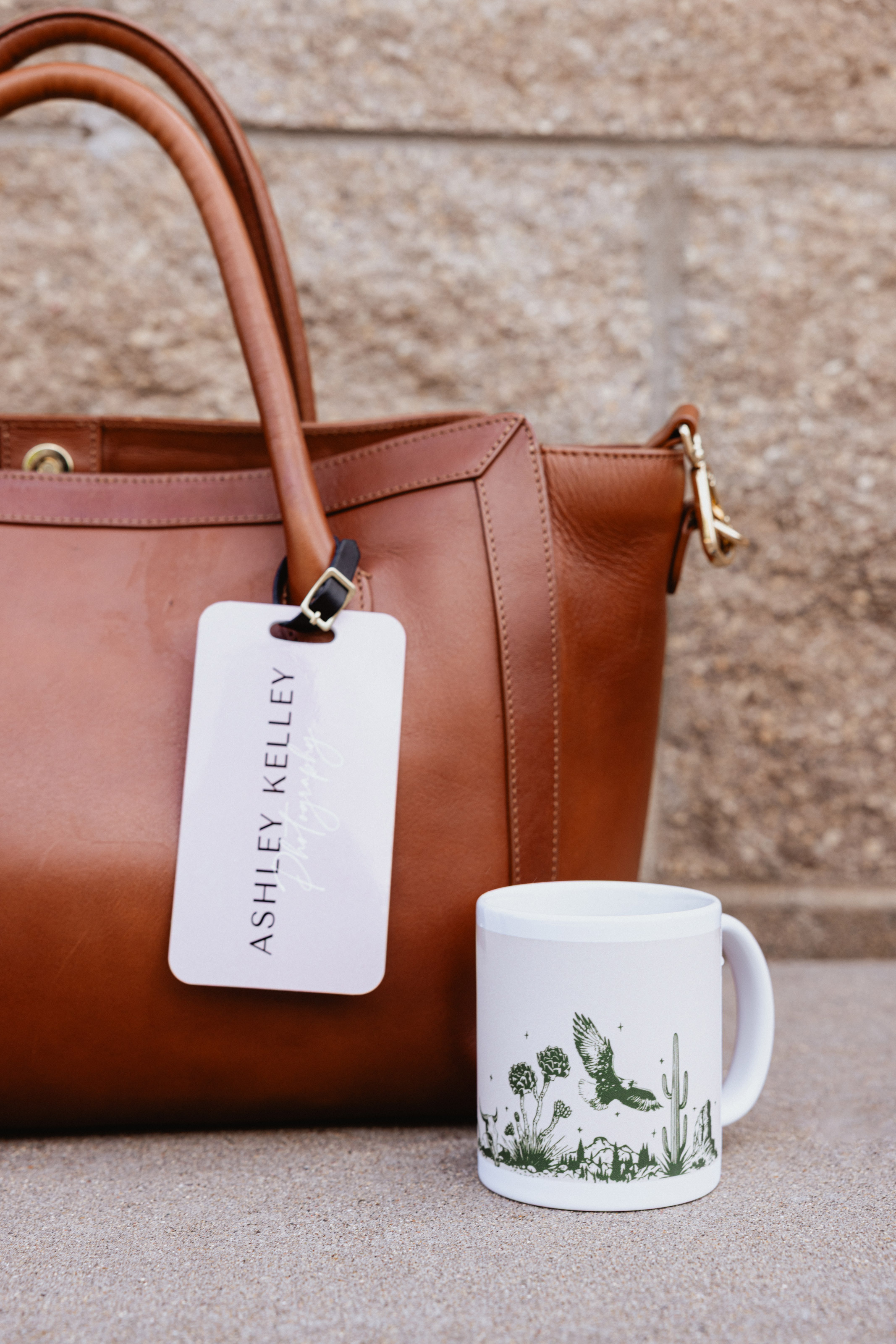 11oz Coffee Mug printed with a nature illustration and sitting outside on the sidewalk by a stone building next to a large brown leather camera bag with a custom-designed Bag Tag displaying the photographer's logo.