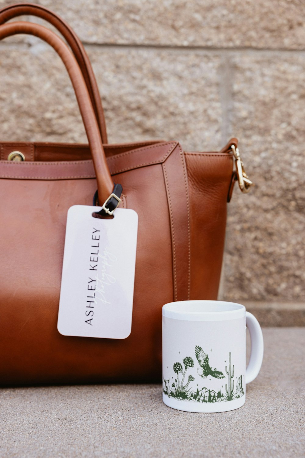 11oz Coffee Mug printed with a nature illustration and sitting outside on the sidewalk by a stone building next to a large brown leather camera bag with a custom-designed Bag Tag displaying the photographer's logo.