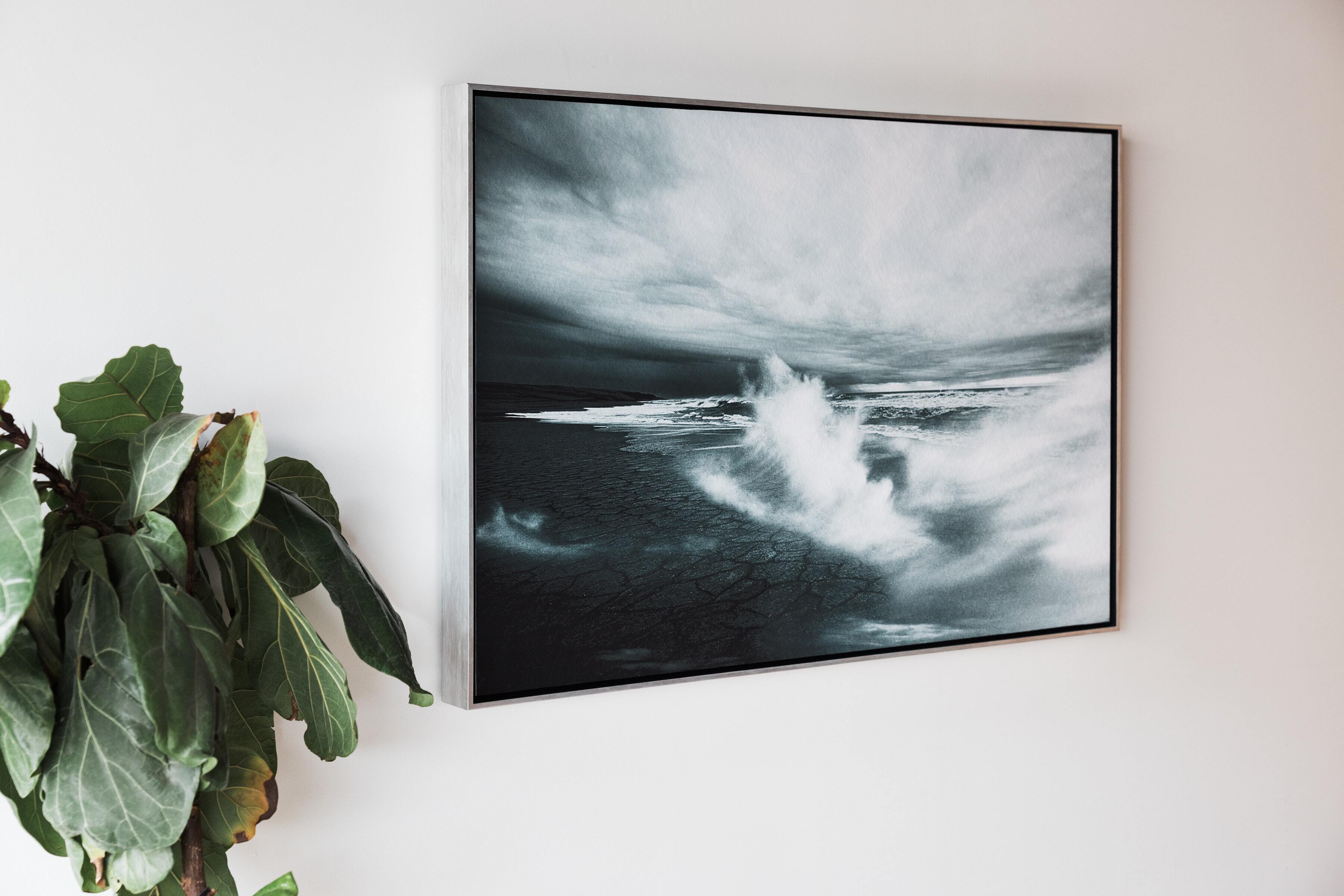 Landscape photograph of dark, stormy ocean waves professionally printed on fine art paper and displayed on a white wall next to a Fiddle Fig plant in a Silver Aged Metallic Float Frame
