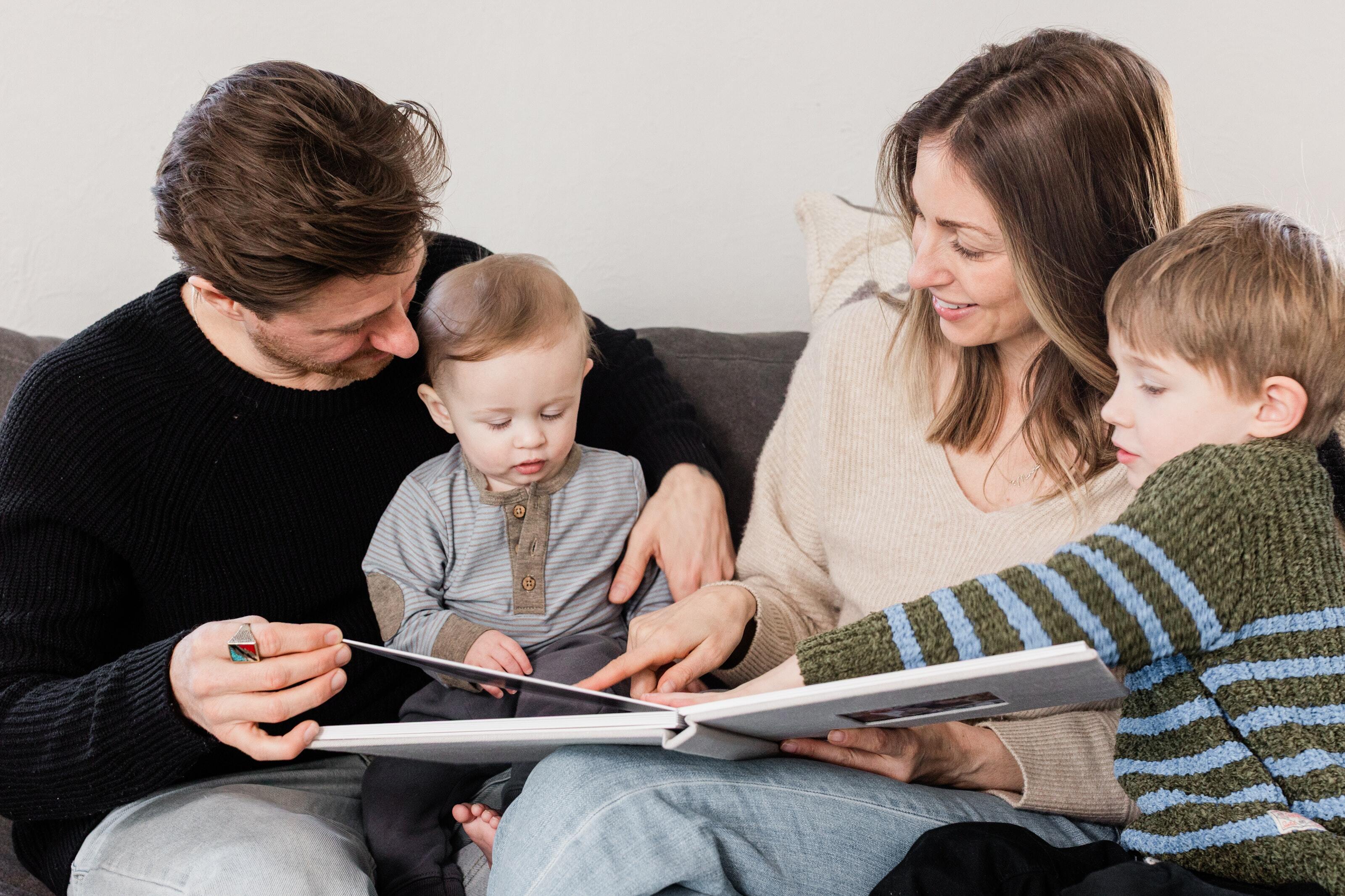 Family on a couch looking through a professional baby photo album together with their toddler and young child.
