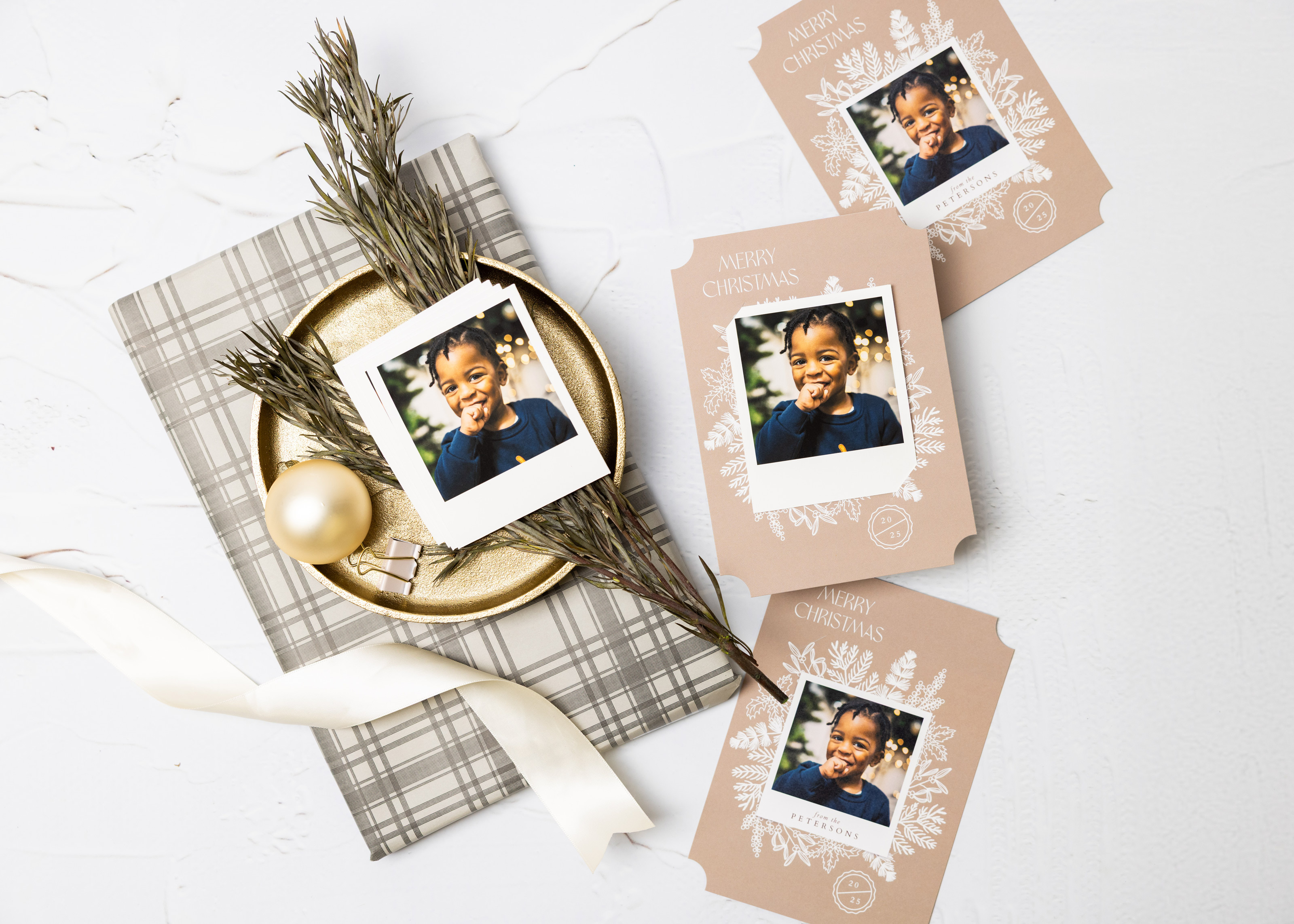 Stack of Mini Snapshots printed with a candid child portrait image next to tan Flat Cards with a "Merry Christmas" message and snapshot prints inserted into slots to hold them to the card. Styled on a white marble surface with holiday decor.