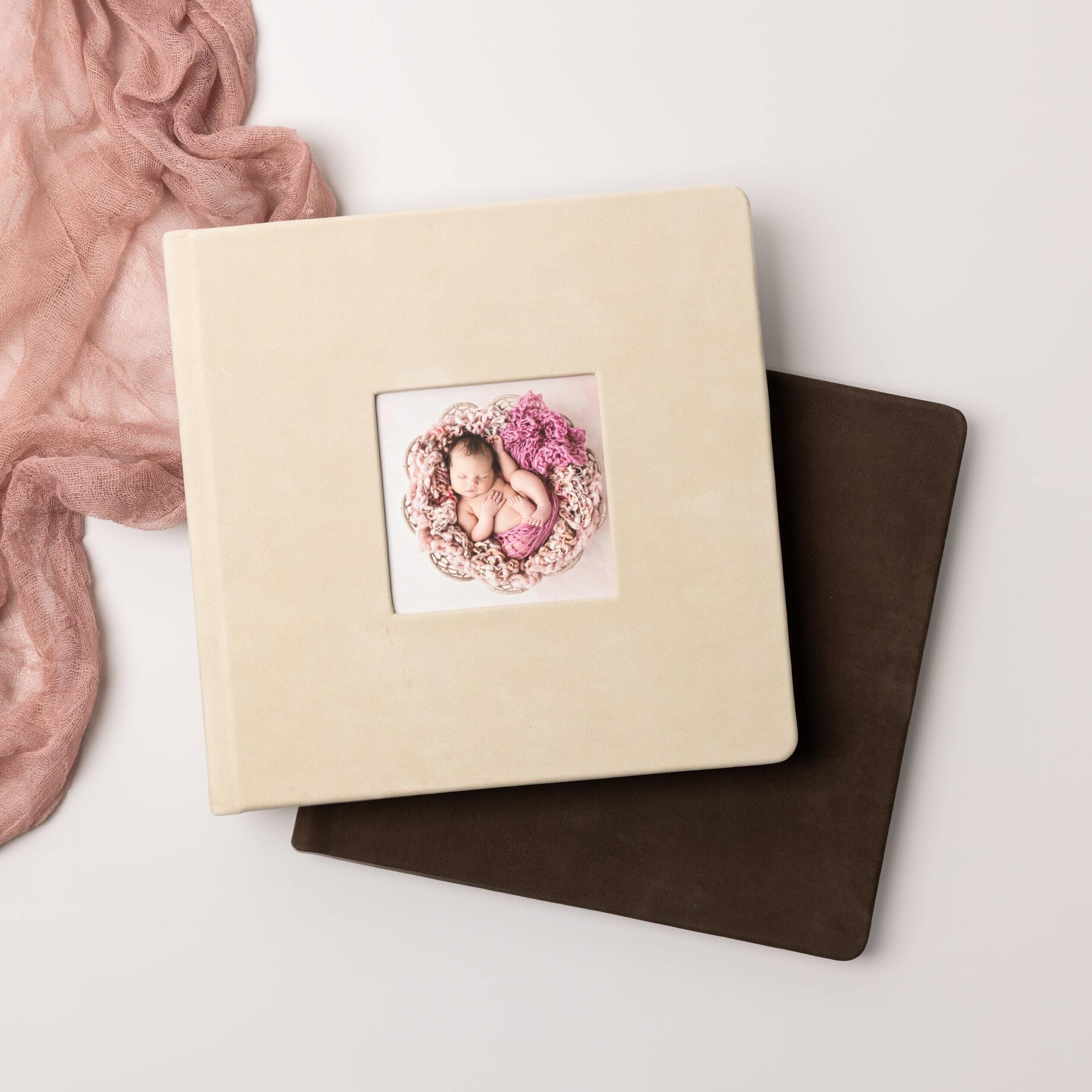 Two professional baby photo albums stacked with a ecru nubuck leather cameo cover featuring a newborn portrait over a espresso nubuck leather album, styled with blush fabric.