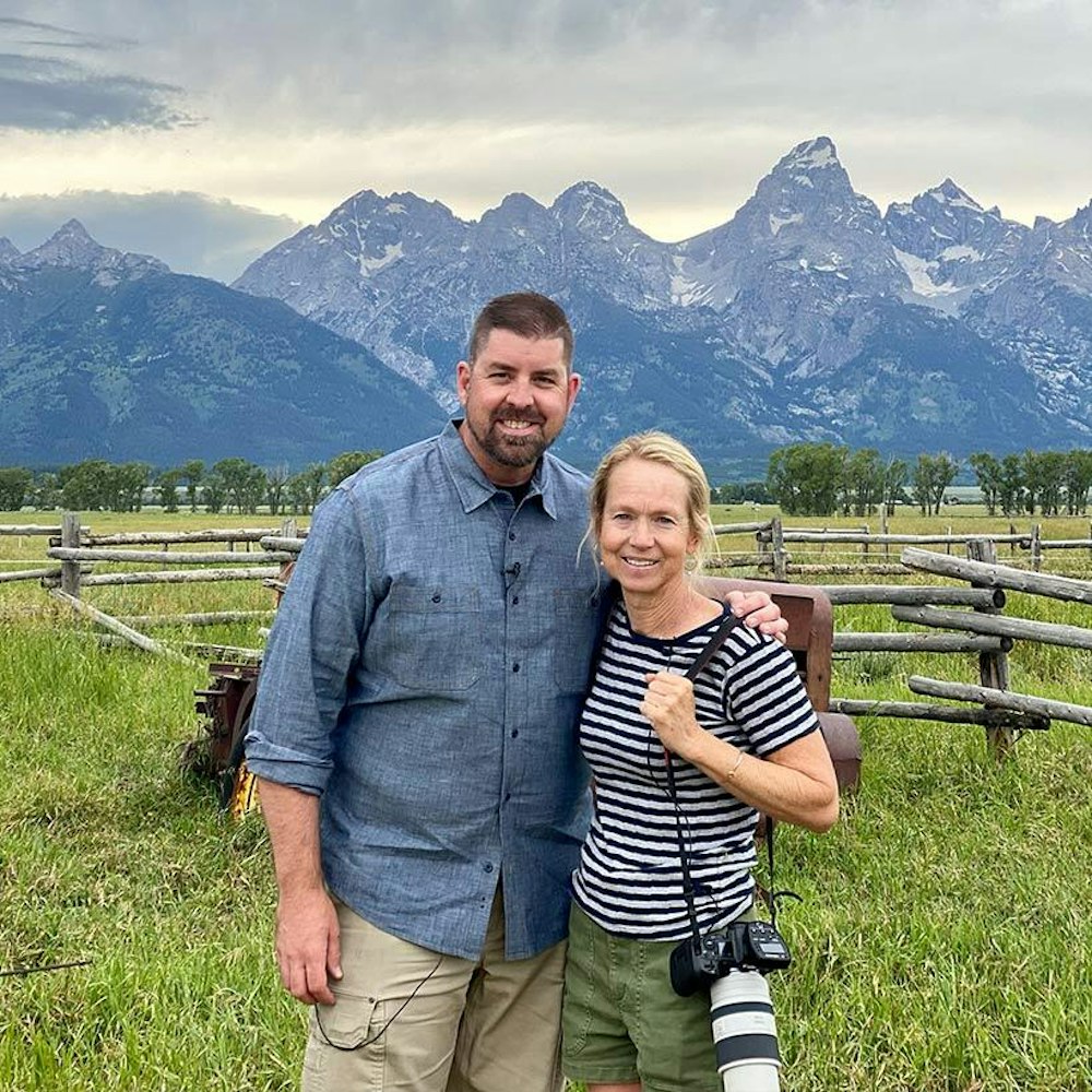 A couple holding a professional camera standing in a grassy field with mountains in the background