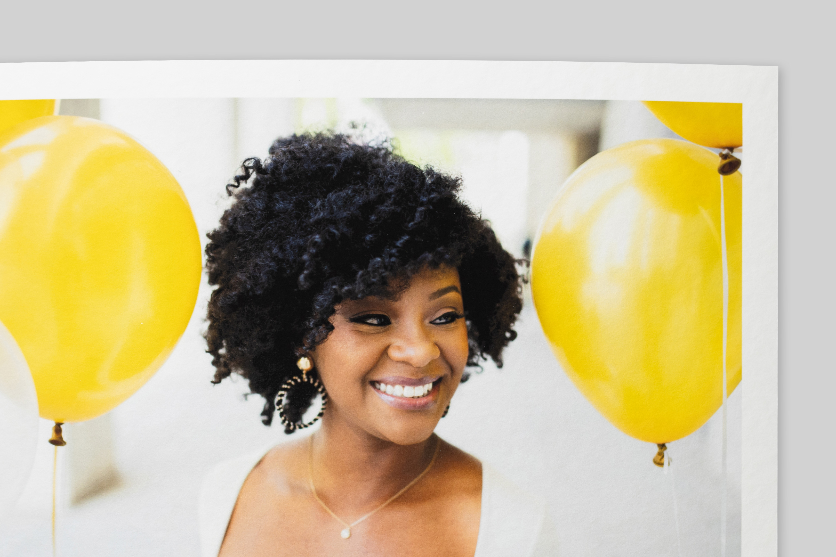 Corner of Torchon Fine Art Print with border, featuring a portrait of a smiling woman with curly hair amid bright yellow balloons. Coarse texture visible on paper surface.