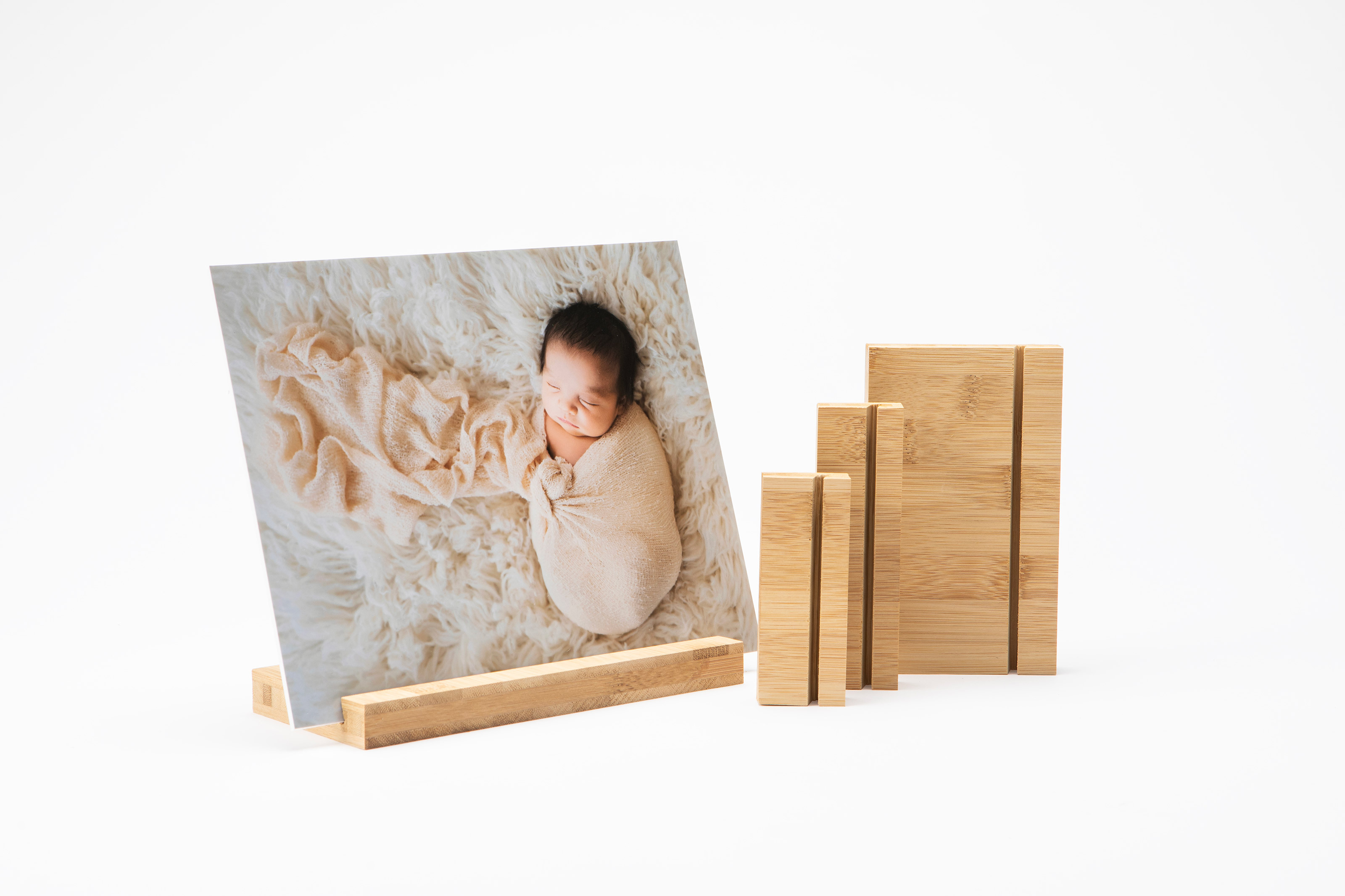 A mounted photo print of a sleeping baby on a bamboo Wood Display Stand next to a set of three stand size options on a plain white background.