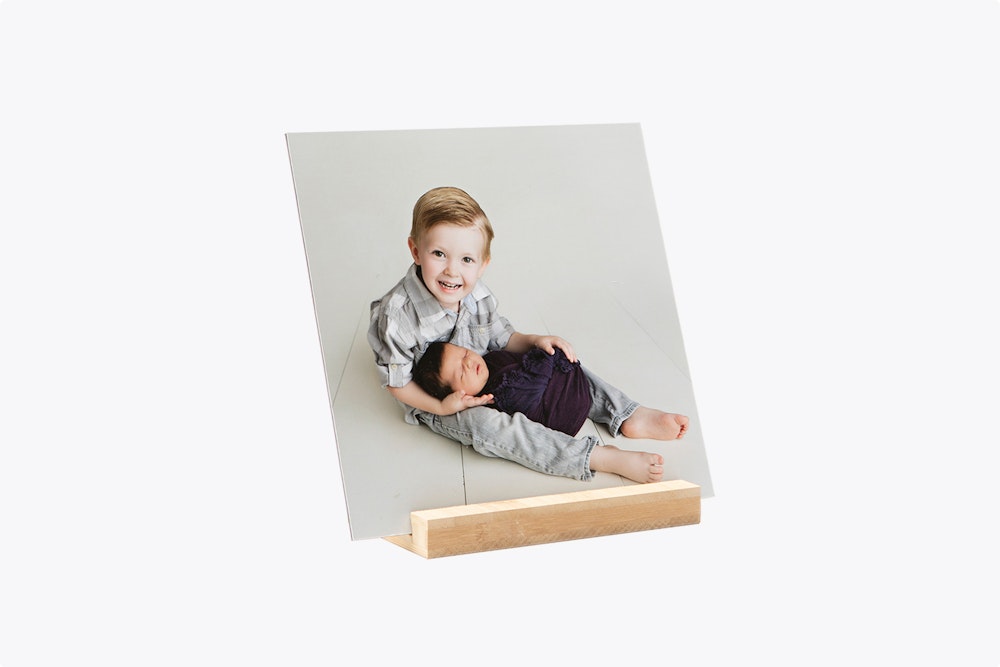 A mounted photographic print on a wooden display stand showing a young boy holding his baby sister on a light gray background