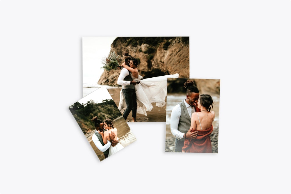 Three printed wedding photographs are arranged on a white surface, featuring a couple embracing on a beach with cliffs in the background.