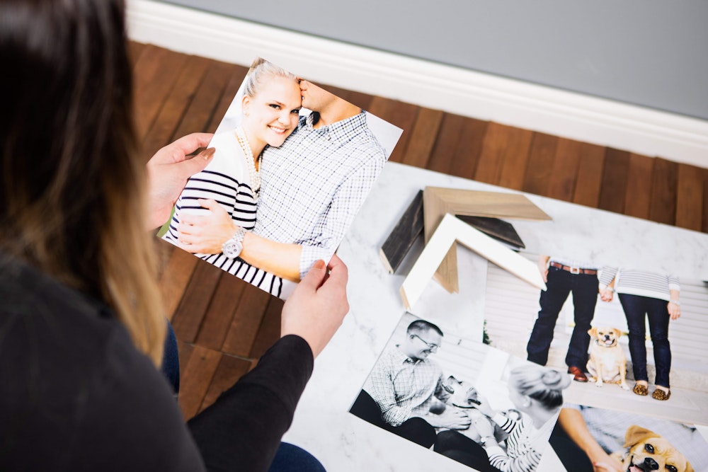 A woman holds a printed photograph of a smiling couple while sitting on the floor surrounded by other printed pictures and frame corner samples.